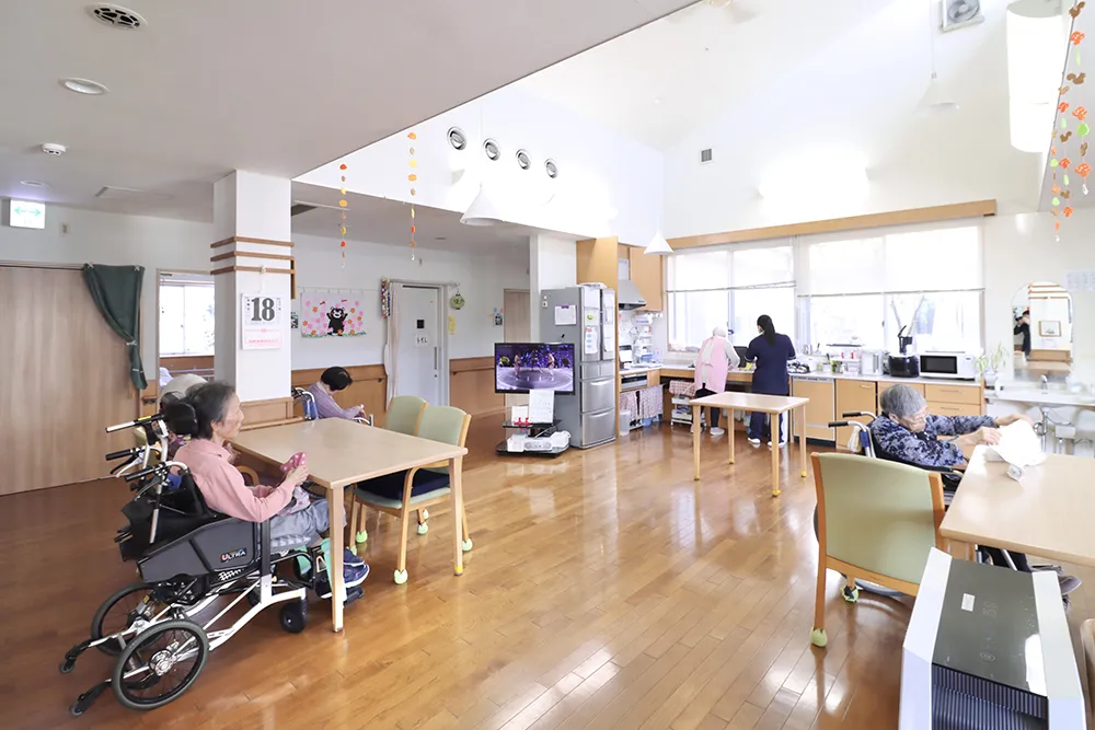 木製家具と大窓を備えた明るい共用スペース。入居者と職員の日常風景。